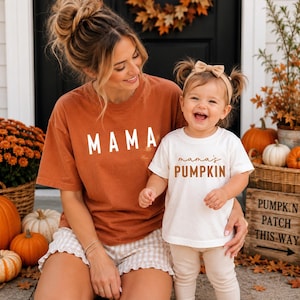 May include: A rust-colored t-shirt with the word MAMA printed in white block letters and a white t-shirt with the text mama's PUMPKIN printed in brown, featuring a short-sleeved design for both garments, worn with light-colored shorts and leggings.