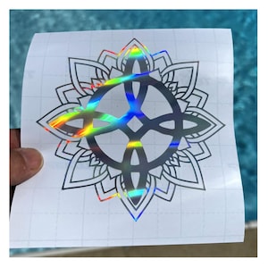 May include: A holographic vinyl sticker featuring a detailed mandala design with a central Celtic knot. The sticker has a rainbow sheen and is held against a blue background. The mandala is surrounded by petal-like shapes.