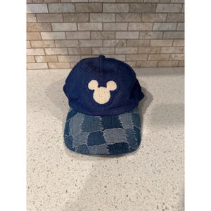 May include: A dark blue denim baseball cap featuring a cream-coloured embroidered Mickey Mouse head silhouette on the front panel. The brim of the cap is constructed from a patchwork of various shades of blue denim fabric in a quilted pattern.