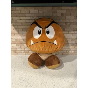 May include: A plush toy shaped like a Goomba character, featuring a round brown body, large white eyes with black pupils, thick black eyebrows, and two small white fangs. The character has two rounded brown feet at the base and a tan underside.