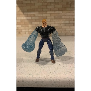 May include: A male action figure with blonde hair, wearing a black short-sleeved shirt and blue pants. The figure features oversized, translucent blue, textured arms that resemble water or ice, with brown shoes and a defined muscular torso.