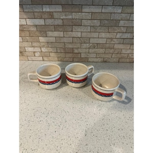 Op de afbeelding: Drie gebroken witte keramische mokken met een gespikkelde afwerking. Elke mok heeft een horizontale decoratieve band rond het midden, bestaande uit een felrode streep omrand door dunne zwarte lijnen en een herhalend donkerblauw halfrond patroon.