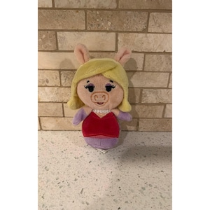 Op de afbeelding: Een knuffel van een varkensfiguur met blond haar, lange wimpers en een roze snuit. Het personage draagt een rode jurk met een witte kraag en paarse mouwen. De knuffel heeft een zachte textuur en is ontworpen met een rond hoofd en oren.