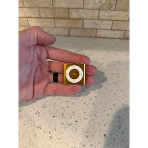 May include: A small, square, gold-colored digital music player featuring a circular white control pad in the center. The control pad includes a play-pause icon and forward and backward track navigation symbols printed in a dark brown color.