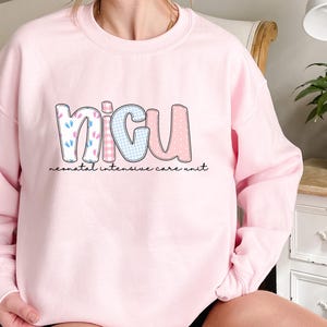 Op de afbeelding: Een lichtroze sweatshirt met de letters "NICU" in een speels lettertype. De letters zijn versierd met patronen zoals babyvoetjes, ruitjes en stippen. De woorden "neonatal intensive care unit" staan eronder gedrukt.