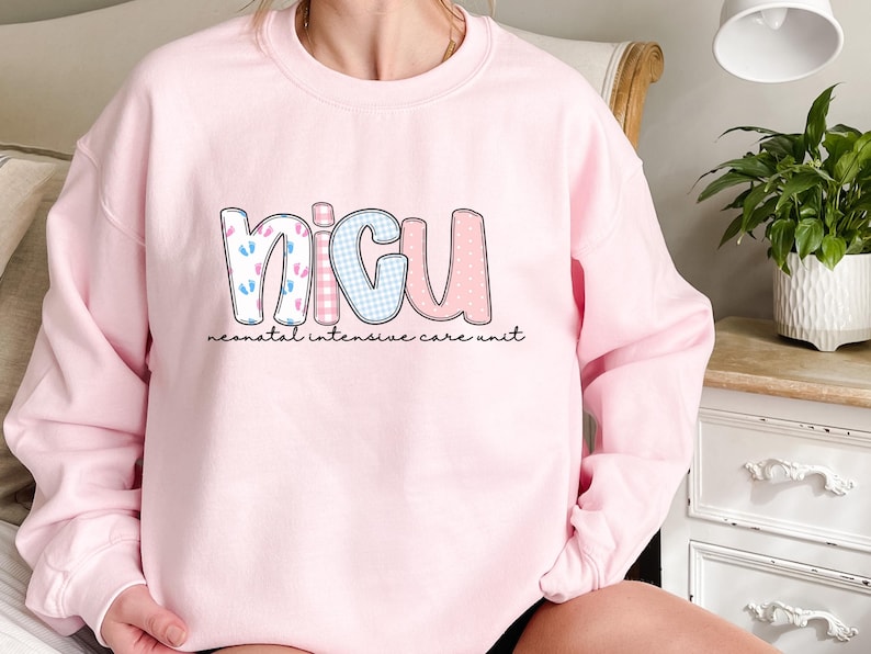 Op de afbeelding: Een lichtroze sweatshirt met de letters "NICU" in een speels lettertype. De letters zijn versierd met patronen zoals babyvoetjes, ruitjes en stippen. De woorden "neonatal intensive care unit" staan eronder gedrukt.
