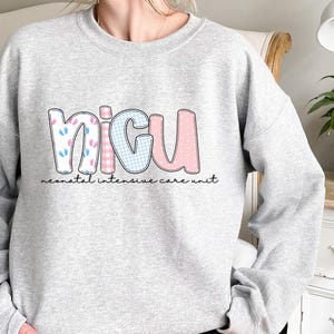 Op de afbeelding: Een lichtgrijs sweatshirt met de letters "NICU" in een kleurrijk ontwerp. De "N" heeft babyvoetjes, de "I" heeft een roze en wit geruit patroon, de "C" heeft een blauw en wit geruit patroon en de "U" heeft een roze stippenpatroon. Daaronder staat de tekst "neonatal intensive care unit".