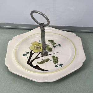 May include: A two-tiered serving tray with a metal handle. The ceramic tray is off-white with a yellow floral design and a yellow border. The handle is silver-colored. The tray has a scalloped edge.