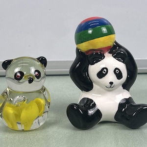 May include: Two panda figurines. One is a clear glass panda with yellow accents. The other is a ceramic panda with black and white coloring, holding a colorful ball. The ball has red, blue, green, and yellow stripes.
