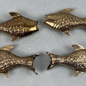 Könnte beinhalten: Vier dekorative Fischfiguren aus Messing. Jeder Fisch hat detaillierte Schuppen, Flossen und ein leicht geöffnetes Maul. Die Fische sind in einem quadratischen Muster angeordnet und zeigen ihre goldene Farbe und ihr aufwendiges Design.