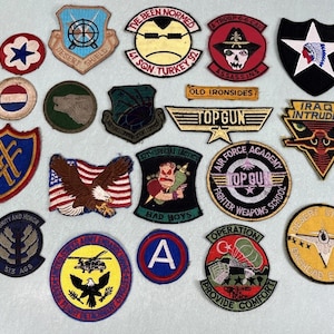 May include: A collection of embroidered military patches with various designs, including eagles, shields, and text like "Top Gun" and "Desert Storm". The patches feature a variety of colors and shapes, showcasing military insignia and unit emblems.