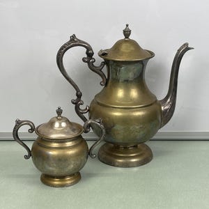 Può includere: Un set da tè in argento placcato composto da teiera e lattiera. La teiera ha un corpo rotondo con un beccuccio curvo e un manico. La lattiera ha un corpo rotondo con un piccolo manico.