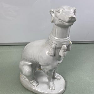 May include: A light gray ceramic dog figurine in a seated position. The dog has a slender build, with a long neck and a detailed face. It wears a collar with decorative tassels. The figurine is on a rounded base.