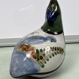 May include: A ceramic duck figurine with a white base, featuring hand-painted details. The duck has a green head, blue beak, and a blue and brown design on its body. The figurine is approximately 4 inches long.