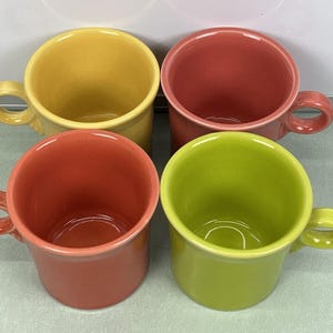 Puede incluir: Cuatro tazas de cerámica coloridas en tonos amarillo, coral, rojo y verde. Cada taza tiene un asa redondeada y una forma cilíndrica. Las tazas están dispuestas en una formación cuadrada, mostrando sus colores vibrantes.