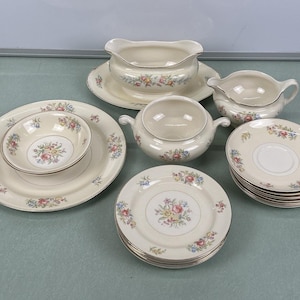Könnte beinhalten: Ein Set aus cremefarbenem Vintage-Porzellan mit floralen Mustern. Das Set umfasst Teller, Untertassen, Schalen, eine Sauciere und eine Servierschüssel mit Deckel. Die Ränder sind goldfarben.