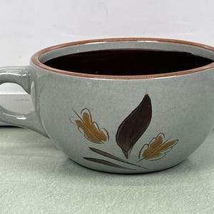 Könnte beinhalten: Eine graue Keramik-Suppentasse mit braunem Rand und Henkel. Die Tasse hat ein Blattdesign in Braun und Gold. Der Innenraum der Tasse ist dunkelbraun.