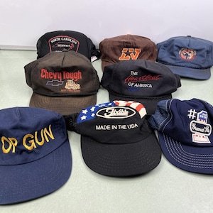 Op de afbeelding: Een verzameling van acht baseballpetten in verschillende kleuren, waaronder marineblauw, bruin en zwart. Verschillende petten hebben geborduurde teksten en logo's, zoals "Top Gun", "Chevy tough" en "Made in the USA". De petten worden gepresenteerd op een lichtgroen oppervlak.