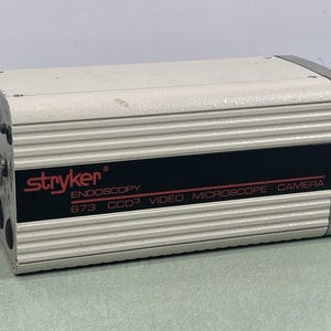Könnte beinhalten: Eine Stryker Endoskopie-Video-Mikroskopkamera, Modell 673 CCD3, in einem rechteckigen, cremefarbenen Metallgehäuse mit einem schwarzen Streifen. Das Stryker-Logo und Produktinformationen sind in Rot und Schwarz gedruckt.