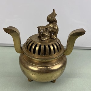Puede incluir: Un quemador de incienso de latón con una figura decorativa de león en la parte superior. El quemador tiene una base redonda, una tapa ranurada y dos asas curvas. El león está detallado con intrincadas tallas.