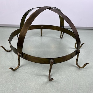May include: A vintage brass pot rack with a circular base and arched supports. The rack features several curved hooks for hanging pots and pans. The metal has a warm, aged patina, suggesting it's an antique kitchen accessory.