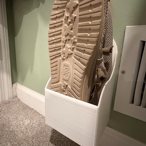 May include: A white plastic shoe holder mounted on a wall with a beige shoe inside.