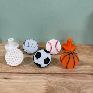 May include: A collection of sports-themed cabinet knobs. The knobs feature designs of a volleyball, soccer ball, baseball, basketball, and other sports equipment. The knobs are white, black, orange, and red.