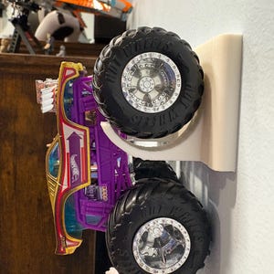 Może przedstawiać: Fioletowo-złoty monster truck Hot Wheels z dużymi czarnymi oponami i srebrnymi felgami. Ciężar&oacute;wka jest zamontowana na białym uchwycie ściennym, prezentując jej design. Widoczne jest logo Hot Wheels.