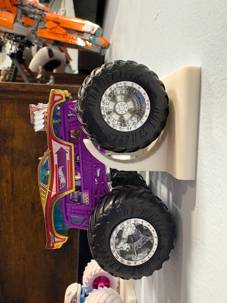 Może przedstawiać: Fioletowo-złoty monster truck Hot Wheels z dużymi czarnymi oponami i srebrnymi felgami. Ciężar&oacute;wka jest zamontowana na białym uchwycie ściennym, prezentując jej design. Widoczne jest logo Hot Wheels.