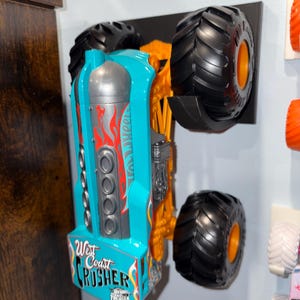 Może przedstawiać: Turkusowo-pomarańczowy monster truck Hot Wheels, "West Coast Crusher", zamontowany na czarnym wyświetlaczu. Samoch&oacute;d ma duże czarne opony z pomarańczowymi felgami i czerwoną grafiką płomieni. Widoczny jest napis "West Coast Crusher".