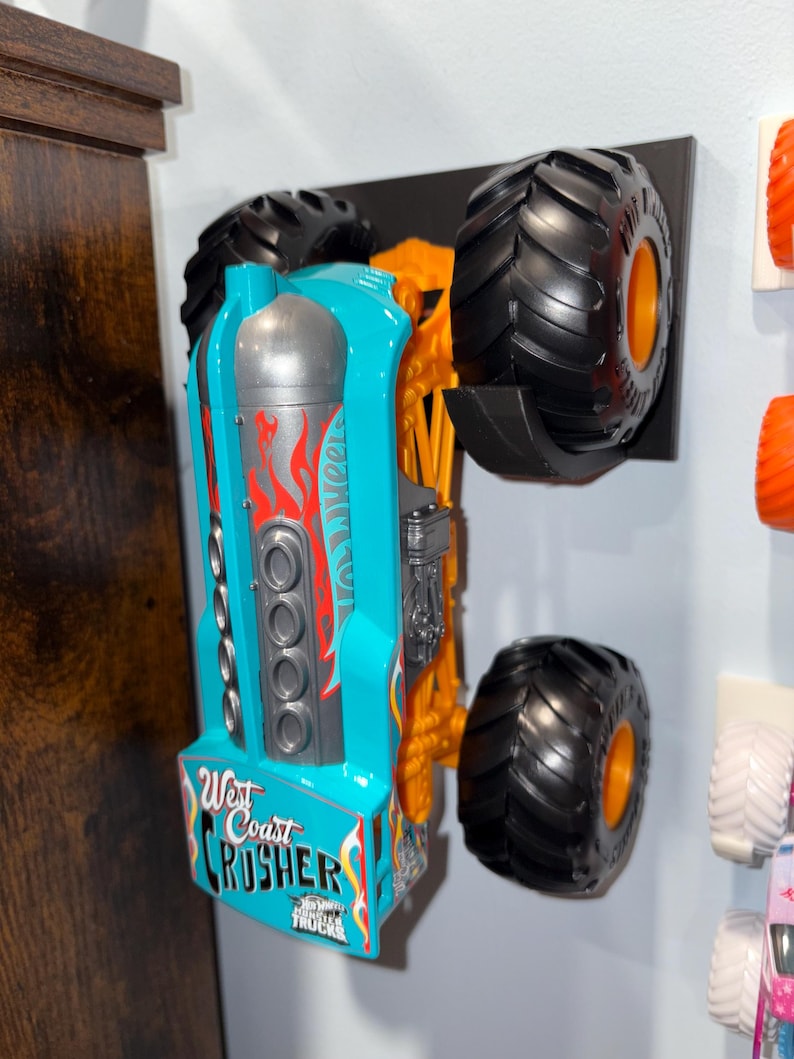Może przedstawiać: Turkusowo-pomarańczowy monster truck Hot Wheels, "West Coast Crusher", zamontowany na czarnym wyświetlaczu. Samoch&oacute;d ma duże czarne opony z pomarańczowymi felgami i czerwoną grafiką płomieni. Widoczny jest napis "West Coast Crusher".