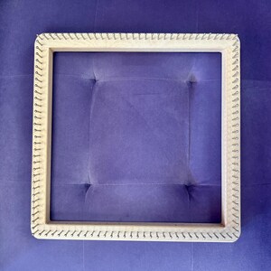 May include: A square wooden knitting loom with metal pegs. The loom is light-coloured and has a square shape. The background is a solid purple colour. The loom is designed for creating knitted items.