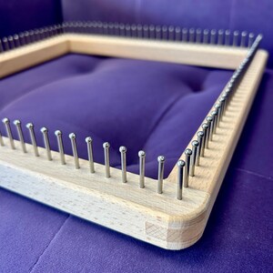 May include: A wooden square knitting loom with evenly spaced metal pegs. The loom is made of light-coloured wood and sits on a purple surface. The pegs are silver and extend upwards from the wood.