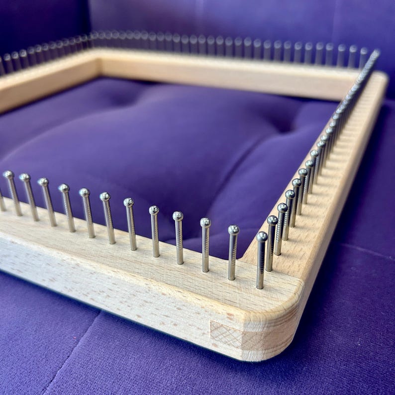 May include: A wooden square knitting loom with evenly spaced metal pegs. The loom is made of light-coloured wood and sits on a purple surface. The pegs are silver and extend upwards from the wood.