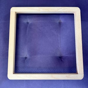 May include: A square wooden frame with rounded corners. The frame is made of light-coloured wood and is empty in the centre. The frame is resting on a purple, tufted surface.
