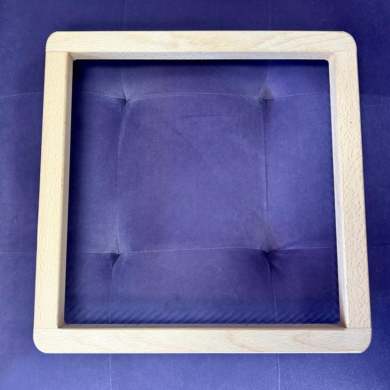 May include: A square wooden frame with rounded corners. The frame is made of light-coloured wood and is empty in the centre. The frame is resting on a purple, tufted surface.