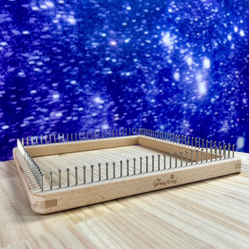 May include: A rectangular wooden knitting loom with metal pegs. The loom has rounded corners and the text "The Yarn Way" is visible on the side. The background is a blue and white starry design.