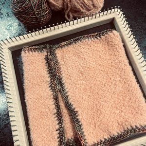 May include: A peach-coloured woven textile is displayed on a wooden loom, with a dark border. Two balls of yarn, one brown and green, and the other peach, are placed near the loom. The textile is likely part of a craft project.