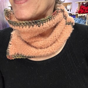 May include: A peach-coloured, fluffy cowl neck warmer with a crocheted edge in shades of green, brown, and black. The neck warmer is made of a soft, textured yarn and is designed to be worn around the neck for warmth and style.