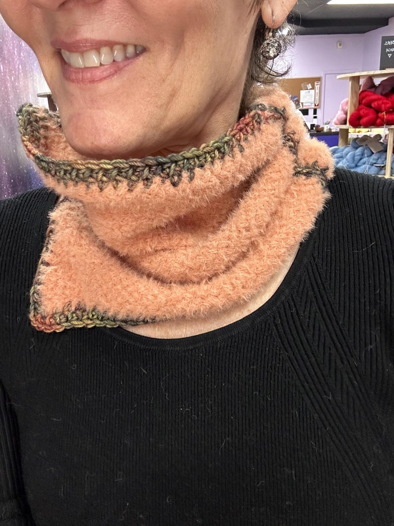 May include: A peach-coloured, fluffy cowl neck warmer with a crocheted edge in shades of green, brown, and black. The neck warmer is made of a soft, textured yarn and is designed to be worn around the neck for warmth and style.