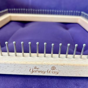 May include: A wooden knitting loom with metal pegs, featuring the text "The Yarn Way." The loom is rectangular with rounded pegs. The background is a solid purple colour.
