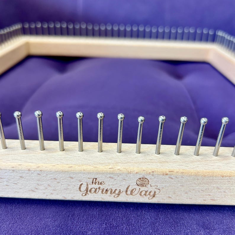 May include: A wooden knitting loom with metal pegs, featuring the text "The Yarn Way." The loom is rectangular with rounded pegs. The background is a solid purple colour.