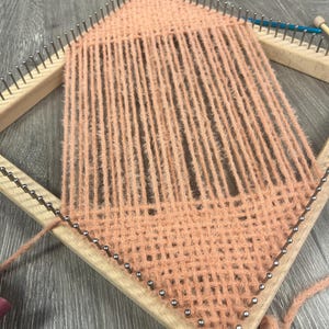 May include: A peach-coloured yarn weaving project in progress on a wooden loom. The loom is diamond-shaped with metal pins holding the yarn in place. The woven section has a textured appearance.