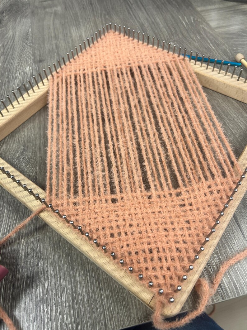 May include: A peach-coloured yarn weaving project in progress on a wooden loom. The loom is diamond-shaped with metal pins holding the yarn in place. The woven section has a textured appearance.