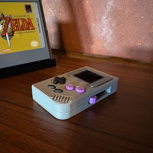 May include: A gray handheld gaming console with black and purple buttons, and a small screen. A game cartridge with the text "The Legend of Zelda: Link's Awakening" is in the background.