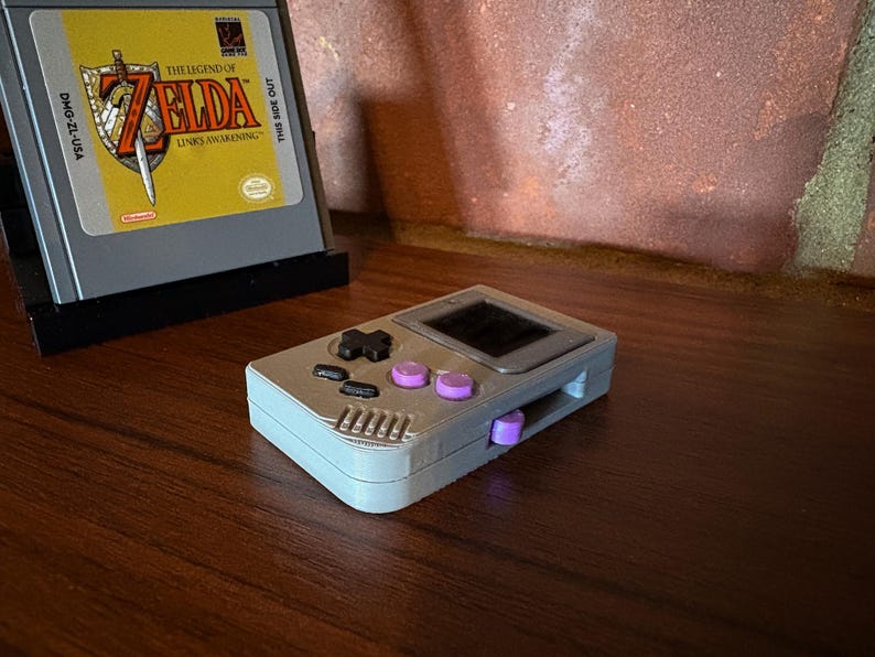 May include: A gray handheld gaming console with black and purple buttons, and a small screen. A game cartridge with the text "The Legend of Zelda: Link's Awakening" is in the background.