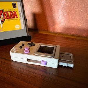 May include: A handheld gaming console with a white and silver finish, featuring purple buttons and a small screen. A game cartridge with "The Legend of Zelda: Link's Awakening" is in the background. The console is on a wooden surface.