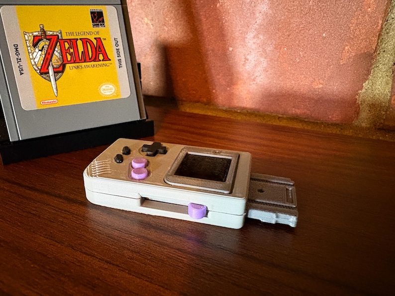 May include: A handheld gaming console with a white and silver finish, featuring purple buttons and a small screen. A game cartridge with "The Legend of Zelda: Link's Awakening" is in the background. The console is on a wooden surface.