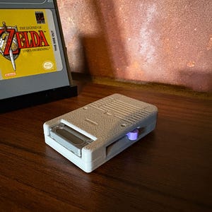May include: A gray handheld game console cartridge adapter with a purple button. The adapter is next to a game cartridge with a yellow label featuring the text "The Legend of Zelda: Link's Awakening". The game cartridge is in a black stand.