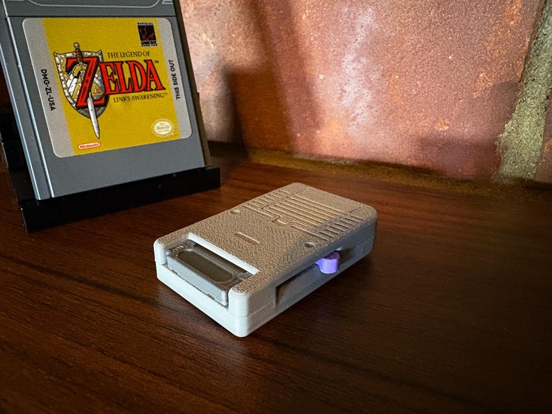 May include: A gray handheld game console cartridge adapter with a purple button. The adapter is next to a game cartridge with a yellow label featuring the text "The Legend of Zelda: Link's Awakening". The game cartridge is in a black stand.